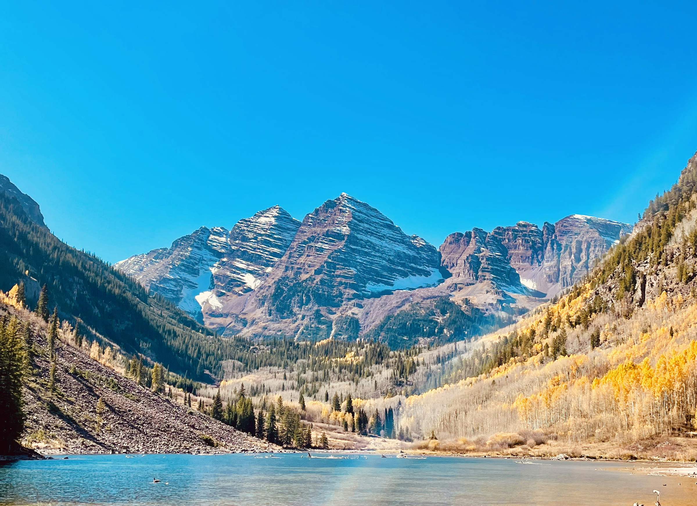 Snow-dusted Maroon Bells above blue lake water and yellow autumn trees