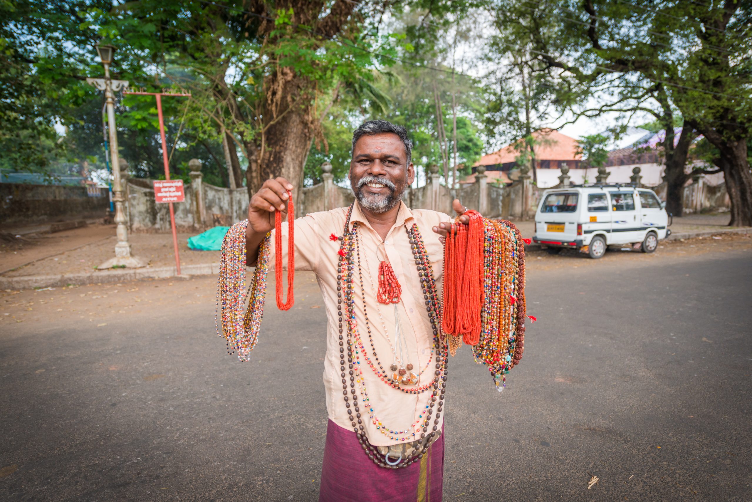 A smiling man holding up strings of red and beige bead necklaces on an open street in soft daylight