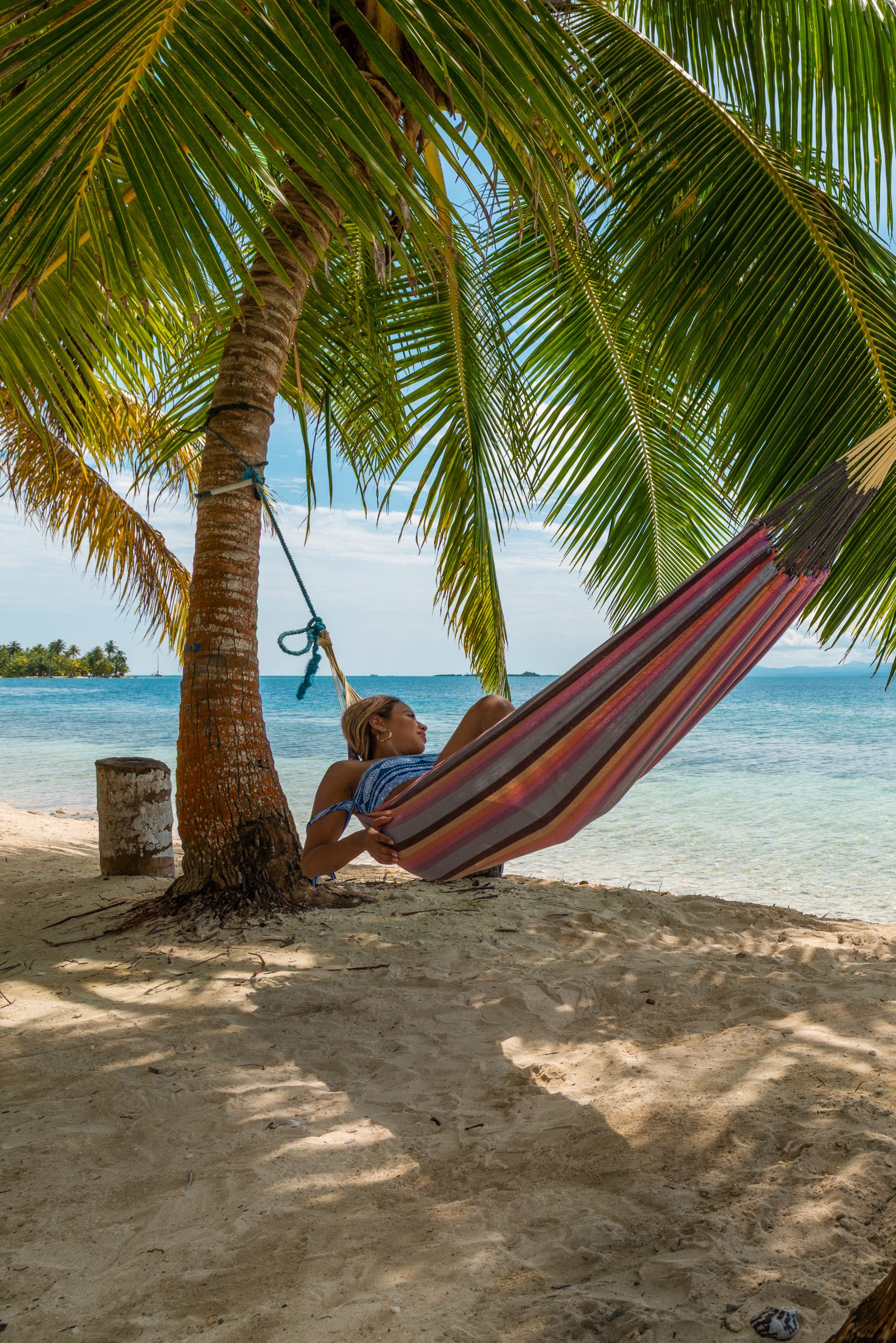 A woman resting in a striped hammock under a palm tree on a beach in Panama, her face in soft open shade