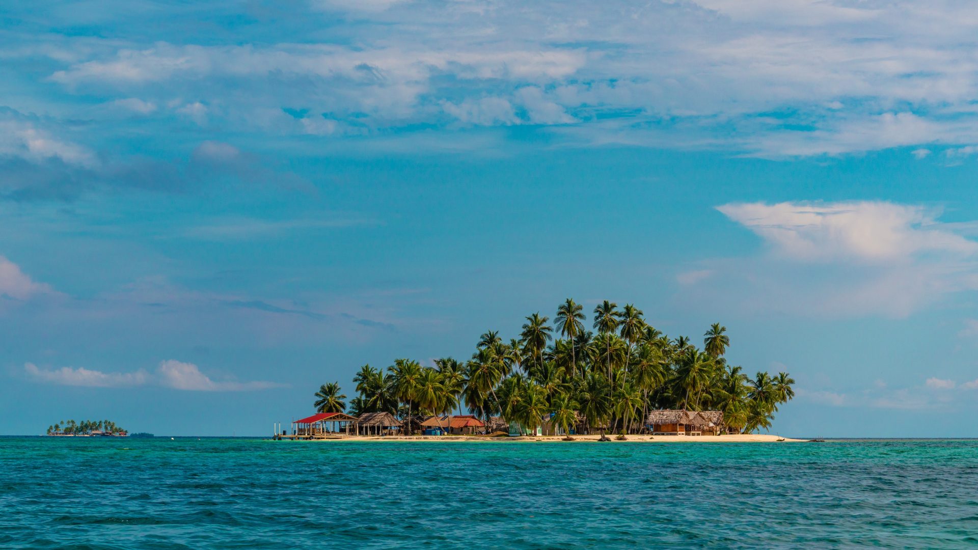 A small palm covered island with thatched huts surrounded by turquoise water under a bright sky