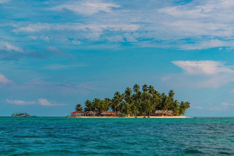 A small palm covered island with thatched huts surrounded by turquoise water under a bright sky
