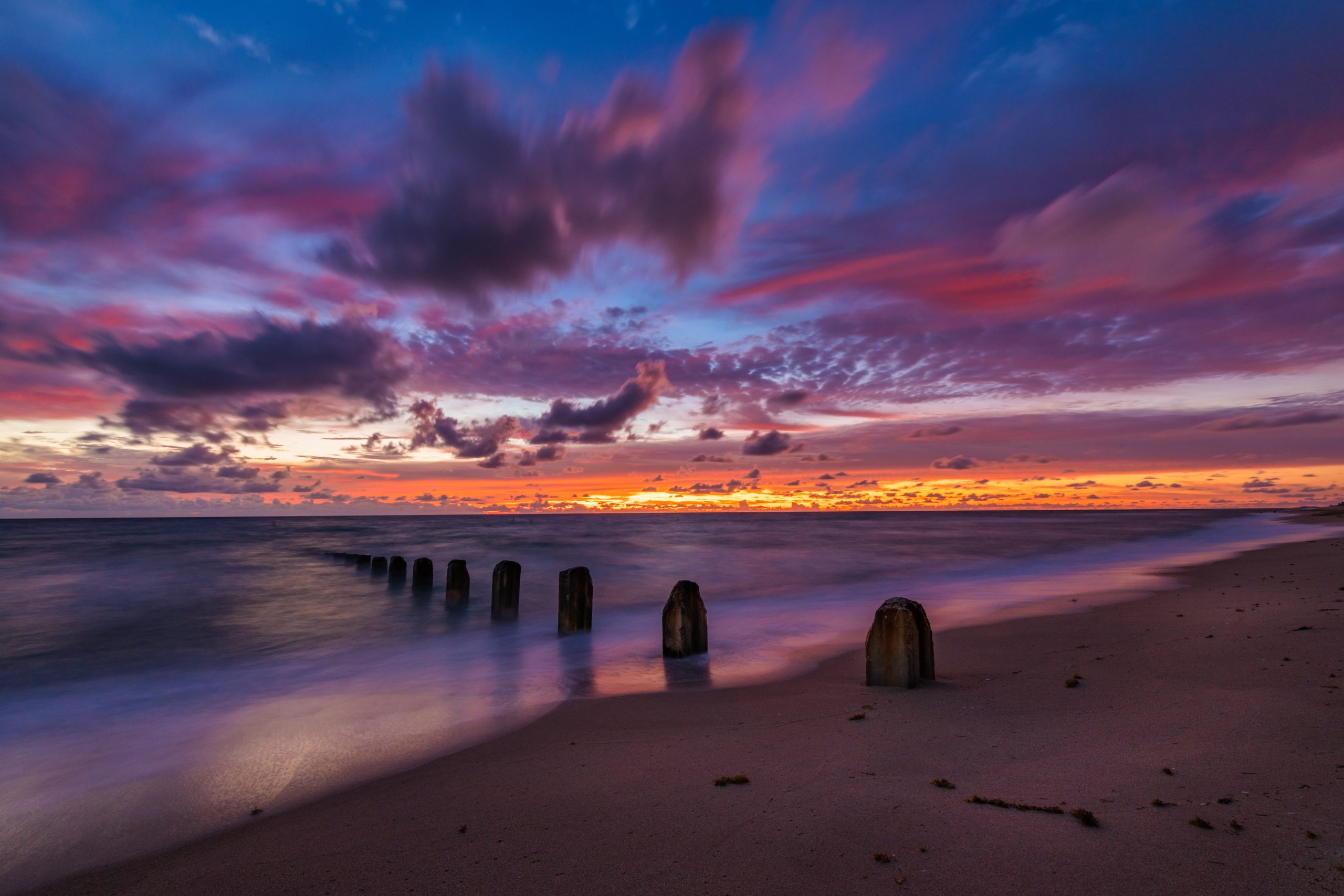 Old posts in the surf under a sky running from orange at the horizon through pink to deep purple