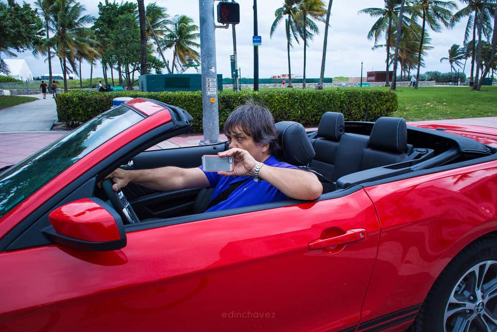 Driver in a red convertible holding up a phone on a palm-lined street