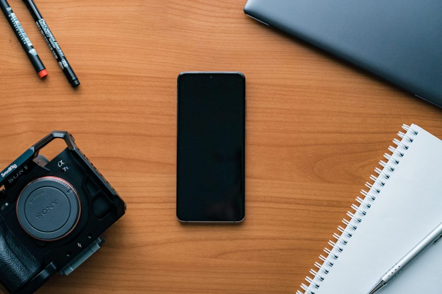 Camera and phone beside a notebook on a wooden desk. (Stock photo, illustrative.)