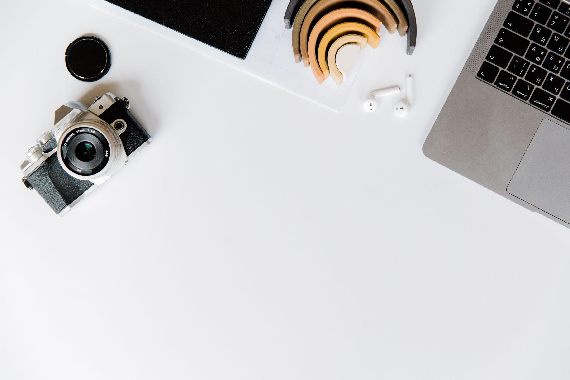 Camera, laptop and earbuds arranged on a white workspace. (Stock photo, illustrative.)