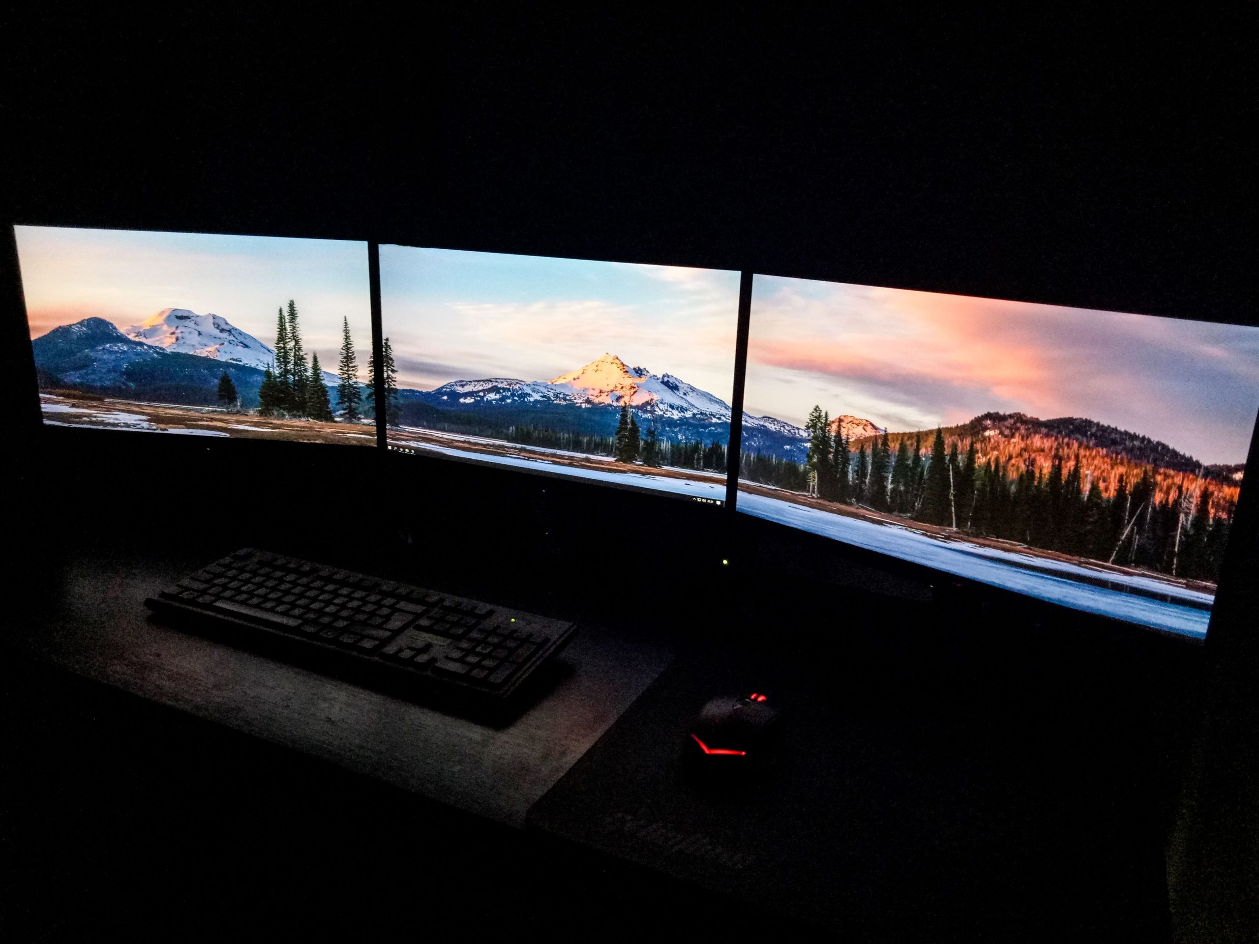 Three monitors displaying a mountain landscape photo in a dark editing room