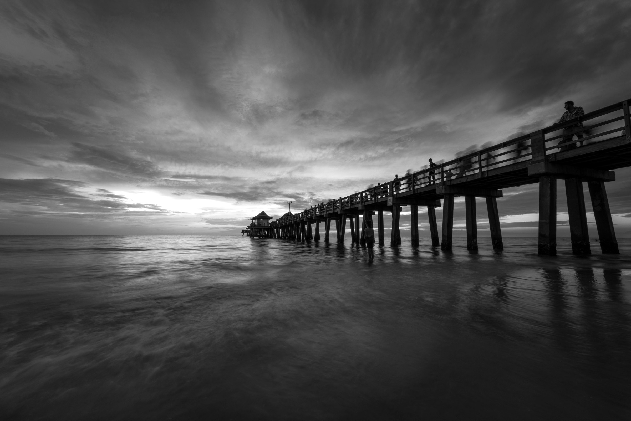 Black and white photograph of the Naples pier at dusk under dramatic layered clouds above rippled water