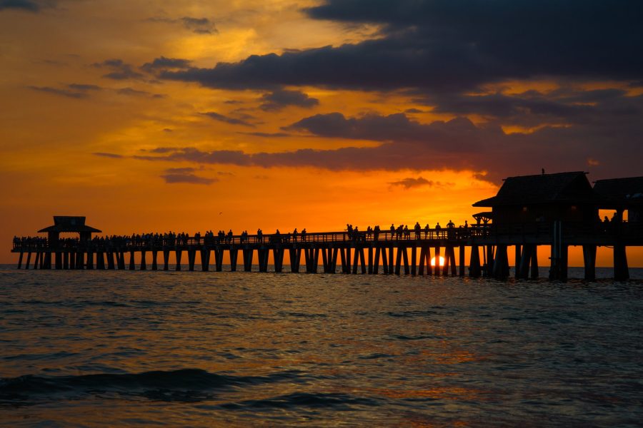 A fiery orange sunset over the Naples pier in Florida, with the sun dropping below the horizon under the pier and the crowd on it in silhouette