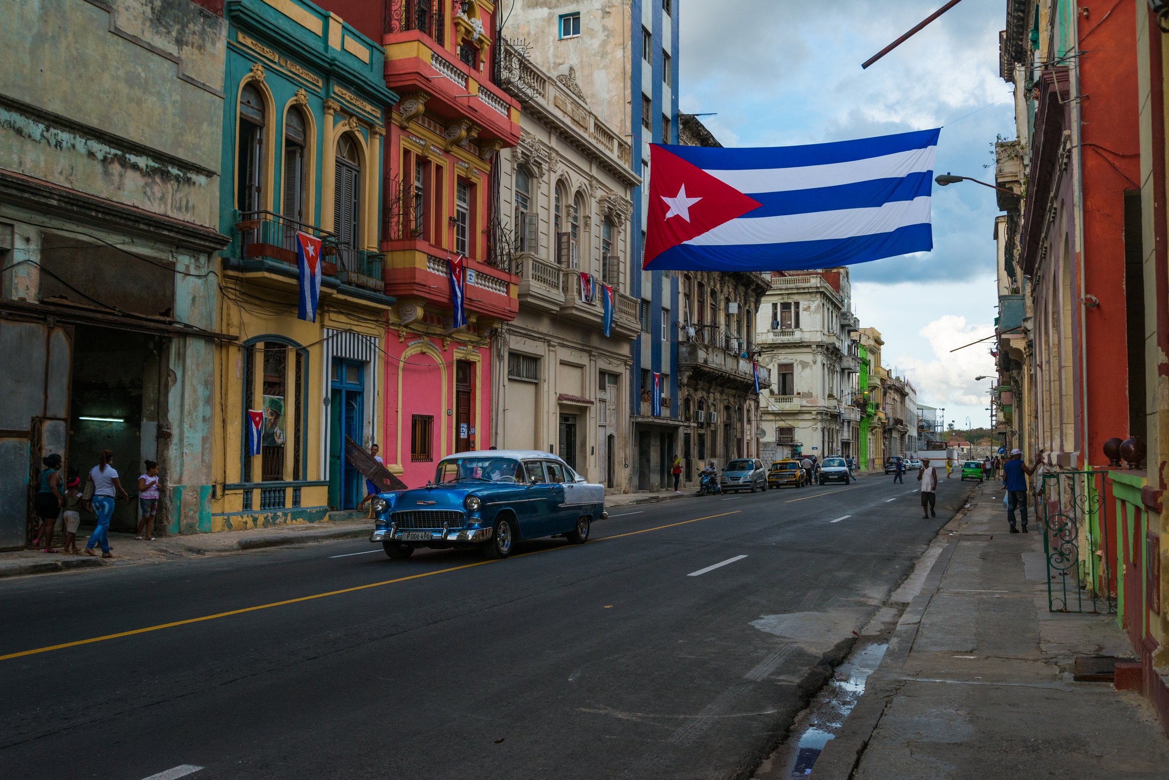 Blue vintage car beneath a large Cuban flag on a colorful street in Old Havana
