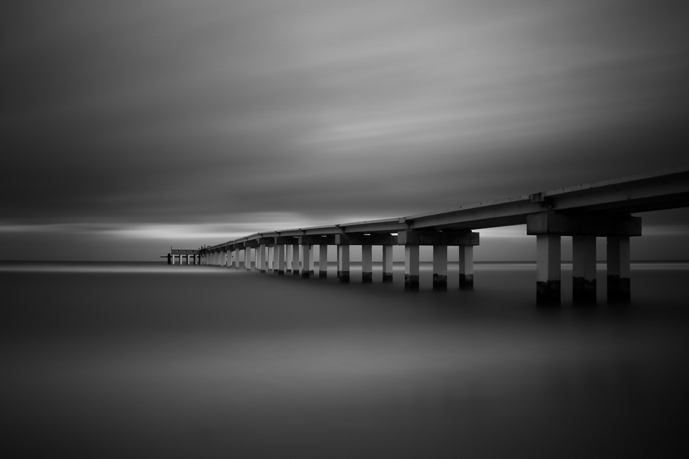 A black-and-white long-exposure of a long concrete pier extending into a calm sea under an overcast sky
