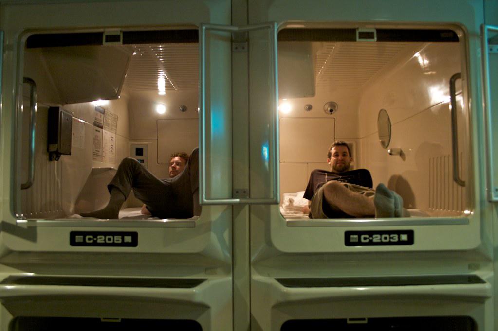 Interior of a capsule hotel in Tokyo, Japan, showing a row of small, futuristic-looking sleeping pods stacked on top of each other. Each pod has a small bed, a TV, and other basic amenities.